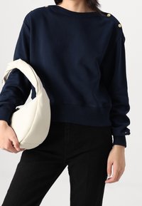 Vrouw draagt een marineblauwe sweatshirt met gouden knopen op de schouders en zwarte broek, terwijl ze een zachte witte handtas onder haar arm houdt.