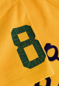Geel katoenen t-shirt met een groene gebroken nummer "8" op de mouw, met een zachte textuur en contrasterende blauwe letters op de voorkant.
