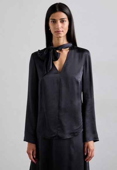 Black satin blouse with a V-neck and long sleeves. Features a large bow tie at the neck and smooth, soft texture. Minimalistic design.