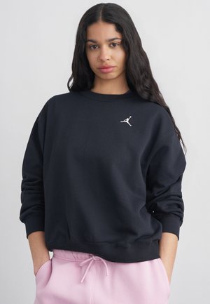 Jeune femme aux longs cheveux foncés portant un sweat-shirt noir avec logo blanc et un pantalon de survêtement rose clair, mains dans les poches, fond neutre.