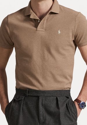 Man in een bruin poloshirt met korte mouwen, ingestopt in een hooggetailleerde, donkergrijze broek met plooien, met handen in de zakken.