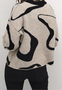 Beige trui met zwart abstract patroon, gemêleerde brei, losse pasvorm en geribbelde zwarte halslijn. Draagt met zwarte broek.