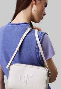 Bílá kožená crossbody taška s embosovaným logem, obdélníkový tvar, nastavitelný popruh a hladká textura, spojená s modrým pleteným topem.