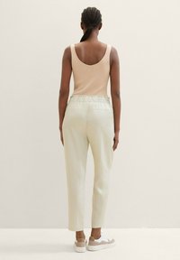 Kvinna med flätat hår som bär en beige ärmlös topp, ljusa krämfärgade byxor med hög midja och vita sneakers, stående mot en neutral bakgrund.