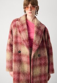 Dubbelzijdig, oversized jas in roze en beige ruitpatroon, gemaakt van zachte, getextureerde wol. Heeft grote revers en zwarte knopen.