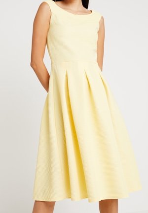 Bezruké světle žluté midi šaty s přiléhavým živůtkem, kulatým výstřihem a plisovanou sukní do áčka, nošené osobou s rukama za zády.