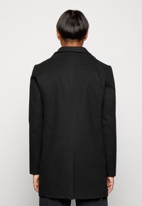 Černý kabát zobrazený zezadu, s hladkou texturou, dlouhými rukávy a strukturovaným límcem. Jednoduché, čisté linie bez ozdob.