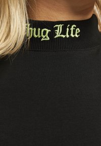 Svart topp med ribbet halslinning og "Thug Life" brodert i lys grønn. Stoffer ser glatte og tettsittende ut.