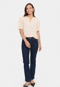 Blusa beige a maniche corte con scollo a V e maniche a palloncino arricciate, abbinata a jeans dritti blu scuro e scarpe nere a punta.