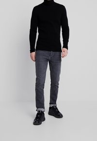 Suéter de cuello alto negro con costillas, jeans ajustados grises, zapatos deportivos negros con detalles en blanco. Posando de pie contra un fondo neutro.