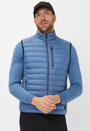 Blue puffer vest with vertical quilted texture, high collar, zippered pockets, and black accents; worn over a blue long-sleeve shirt.
