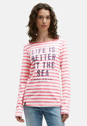 Junge Frau mit lockigem Haar, die ein rosa-weiß gestreiftes Langarmshirt mit dem Text "Das Leben ist besser am Meer" und eine blaue Jeans trägt.