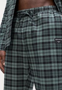 Πράσινα και μαύρα καρό πιτζάμα με ελαστική μέση και πλαϊνές τσέπες, κατασκευασμένα από μαλακό ύφασμα; διαθέτει λογότυπο στο πλάι.