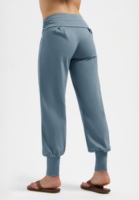 Person, der hochgeschnittene, lockere blaue Jogginghosen mit umgeschlagenen Knöcheln und braunen Slippern trägt, von hinten gezeigt.