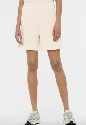 Weibliches Model trägt beige Shorts mit hoher Taille und umgeschlagenen Säumen sowie weiße Turnschuhe, steht vor einem einfarbigen Hintergrund.