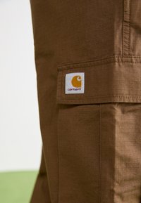 Braune Carhartt-Hosen bestehen aus einem strukturierten Stoff mit einem ausgeprägten Karomuster, einer aufgesetzten Tasche und einem Logopatch in gelb und weiß.