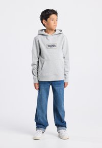 Sudadera gris con capucha, bolsillo frontal y parche de logo, combinada con jeans azules de corte holgado y zapatillas blancas.