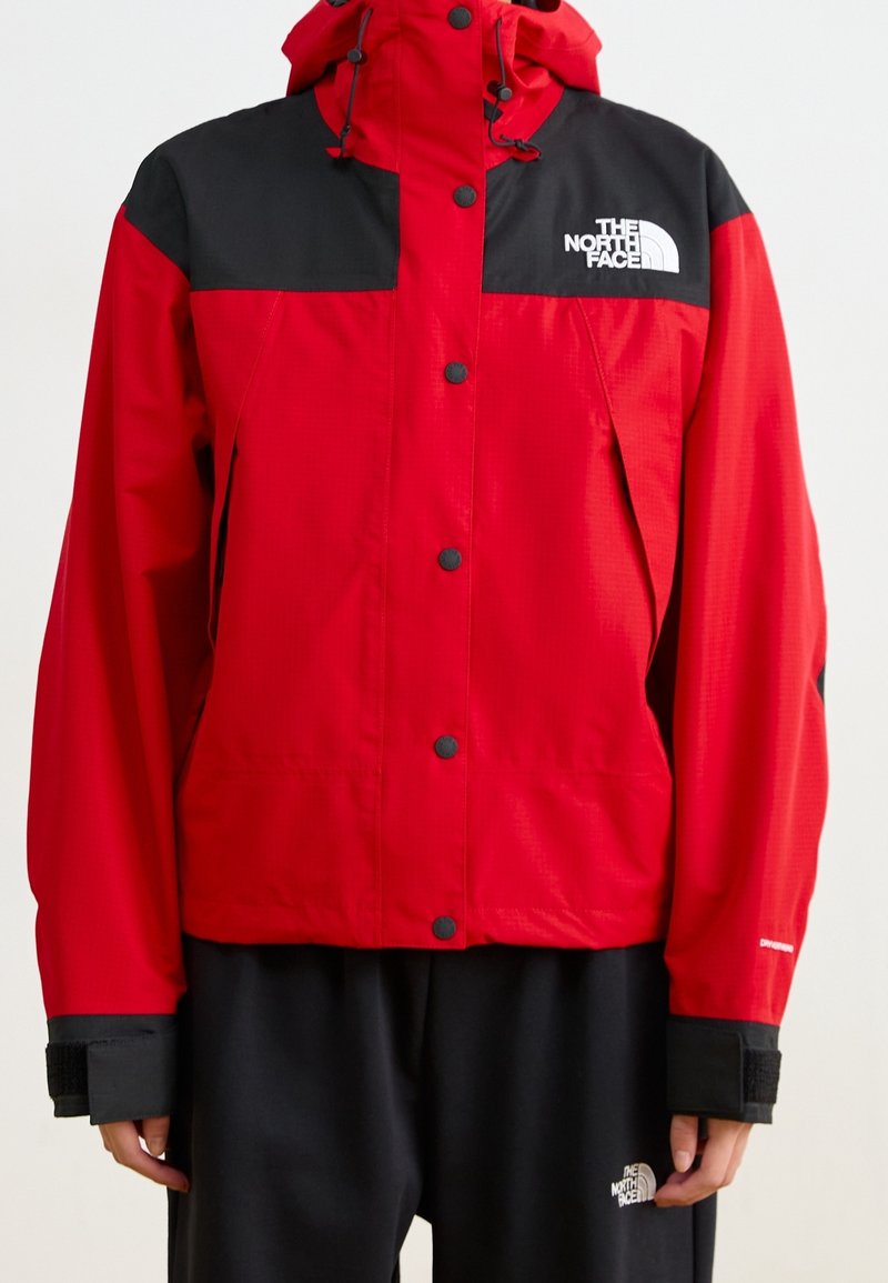 Rote und schwarze Windjacke mit Kapuze, Druckknöpfen, zwei Seitentaschen und Logo auf der Brust und am Ärmel.