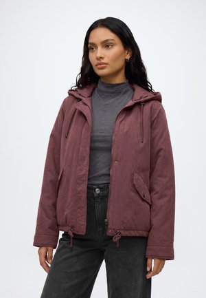 Femme portant une veste à capuche violette par-dessus un col roulé gris avec un pantalon noir, debout devant un fond clair uni.
