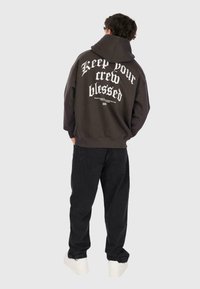 Person in einem dunklen Hoodie mit dem Text "Halte deine Crew gesegnet" auf dem Rücken, schwarzen Hosen und weißen Sneakers, steht mit dem Rücken zur Kamera.