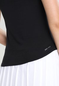 Primer plano de una persona que lleva una camiseta sin mangas negra Dri-Fit y una falda blanca con pliegues, mostrando la parte superior del torso y el brazo.