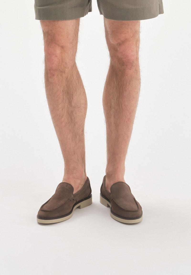 Brązowe zamszowe buty slip-on o stonowanej, teksturowanej powierzchni i kontrastowej beżowej gumowej podeszwie, umieszczone z przodu odkrytych nóg.