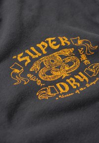 Czarny t-shirt z materiału z textured pomarańczowym wzorem graficznym, stylizowanym tekstem i emblematycznym motywem węża na środku.