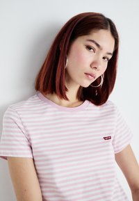 Rosa og hvit stripete t-skjorte, kort ermer, rund hals, med en liten rød Levi's-logo på venstre side. Mykt bomullsstoff.
