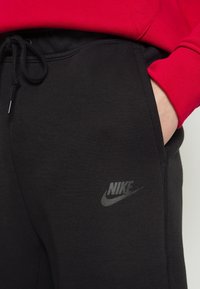 Zwarte trainingsbroek met een trekkoord in de taille, gemaakt van zacht materiaal. Heeft een subtiele grijze Nike-logo aan de zijkant en een rode sweatshirt erboven.