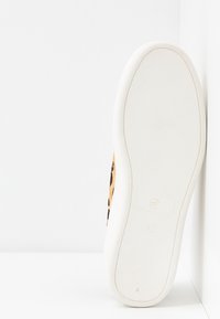 Semelle de chaussure en caoutchouc blanc avec une fine texture de crampons, présentée verticalement sur un fond blanc, révélant partiellement un côté imprimé léopard.