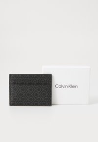 Portafoglio in pelle nera con motivo in rilievo del logo CK, dotato di slot su entrambi i lati. Accompagnato da una scatola bianca di Calvin Klein.