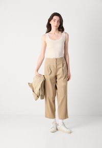 Beige tanktop, hoge taille kaki broek en witte sneakers. Model houdt een bijpassende beige jas vast. Soepele texturen en moderne pasvorm.