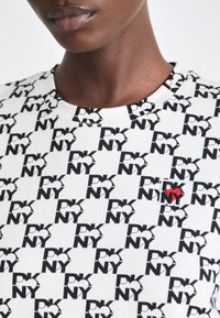 Jasni prikaz osobe koja nosi bijelu majicu s ponavljajućim crnim "DKNY" logotipima i jednim logotipom s crvenim srcem.