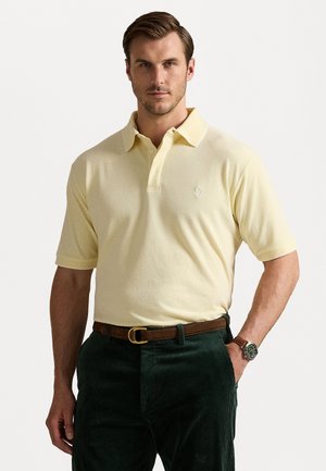 Kollane polo särk lühikeste varrukatega, kaelus ümberkaudse disainiga, rinna peal on diskreetne logo, koos tumeroheliste sametpükste ja vööga.