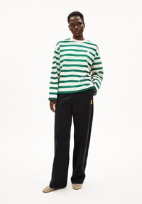 Frau steht mit einem grün-weiß gestreiften Langarmshirt, schwarzen weit geschnittenen Hosen mit gelbem Logo und beigefarbenen Schuhen vor einem weißen Hintergrund.