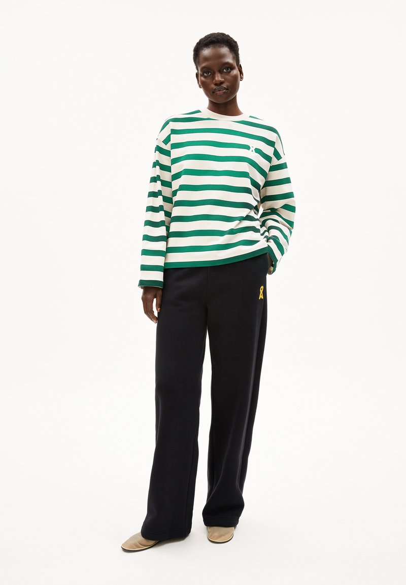 Frau steht mit einem grün-weiß gestreiften Langarmshirt, schwarzen weit geschnittenen Hosen mit gelbem Logo und beigefarbenen Schuhen vor einem weißen Hintergrund.