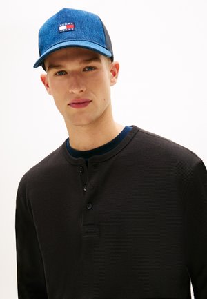 Juoda ilgomis rankovėmis Henley stiliaus marškinėliai iš tekstūruoto audinio su trimis sagomis, derinami su mėlyna džinsine kepure su logotipo lipduku.