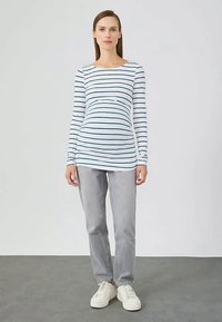 Egy nő áll, aki fehér, hosszú ujjú felsőt visel sötét vízszintes csíkokkal, világosszürke farmert és fehér sportcipőt hord a szürke padlón.