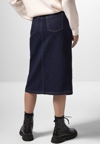 Falda de mezclilla oscura hasta la rodilla con una cintura ajustada, dos bolsillos traseros y detalles de costura amarilla. Combinada con botas negro con cordones.