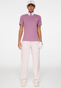 Golfpolo i lavendel med hvid krave og tre-knaps lukning, kombineret med lyserøde bukser. Modellen bærer en kasket og handske.