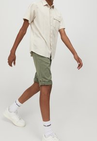 Lichtbeige overhemd met korte mouwen en een borstzak, gecombineerd met opgerolde olijfgroene shorts en witte sneakers en sokken.