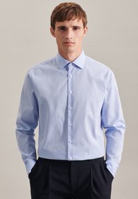 Camicia azzurro chiaro con un piccolo motivo a cerniera. Caratterizzata da un colletto a punta, maniche lunghe e una vestibilità sartoriale. Indossata con pantaloni scuri.