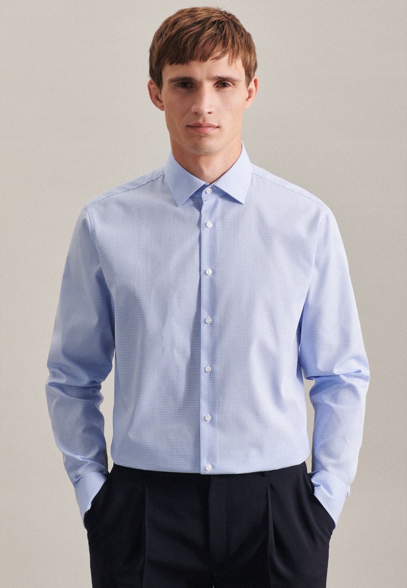 Camicia azzurro chiaro con un piccolo motivo a cerniera. Caratterizzata da un colletto a punta, maniche lunghe e una vestibilità sartoriale. Indossata con pantaloni scuri.