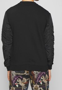 Sudadera negra con mangas negras texturizadas, escote redondo y puños acanalados. Pantalones estampados con diseños intrincados visibles en la parte inferior.