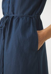 Hand liegt in der seitlichen Tasche eines navyblauen Kleides mit Kordelzug in der Taille und geknöpftem Vorderteil.