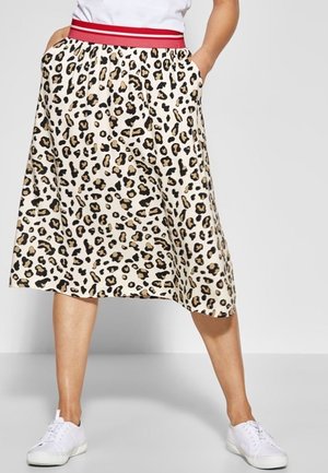 Femme portant une jupe léopard longueur genou avec une ceinture élastique rayée rouge, des baskets blanches, les mains dans les poches de la jupe.