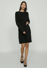 Γυναίκα με μαύρη μακρυμάνικη φόρμα και ζώνη στη μέση, φορώντας μαύρες γόβες με λουριά, στέκεται μπροστά από έναν απλό φωτεινό φόντο.