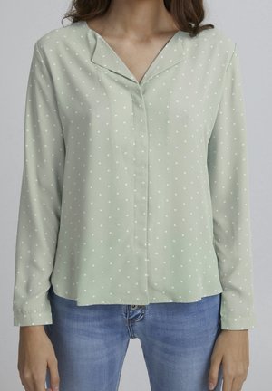 Femme portant un chemisier vert clair à manches longues avec des pois blancs et un jean bleu, debout devant un fond uni.