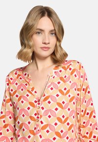Femme aux cheveux blonds mi-longs portant un chemisier à motifs rose, orange et beige avec des boutons et un col en V.