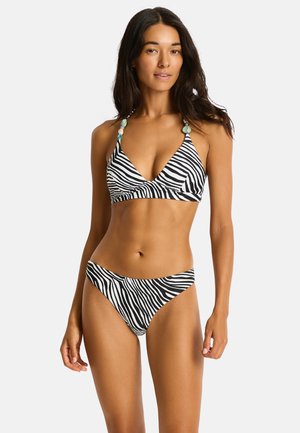 Vrouw draagt een zwart-wit zebraprint bikini met haltertop met decoratieve kralen, staand voor een effen lichte achtergrond.
